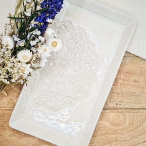 Könnte beinhalten: Ein rechteckiges Tablett aus weißer Keramik mit einem erhabenen, geprägten Mandala-Design. Das Tablett hat einen leicht erhöhten Rand und eine glatte, glänzende Oberfläche.
