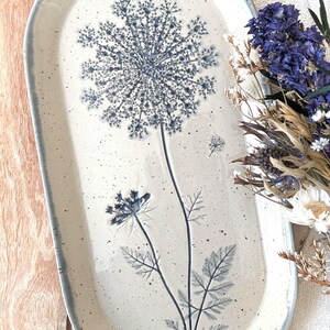 Könnte beinhalten: Ein ovales Serviertablett aus Keramik mit gesprenkelter cremefarbener Glasur und einer blauen botanischen Illustration einer wilden Möhrenblüte. Das Tablett hat einen erhöhten Rand mit blauer Kante und zeigt einen detaillierten Blütenstängel.