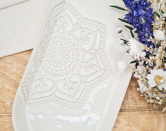 Könnte beinhalten: Ein weißes, ovales Tablett aus Keramik mit einem erhabenen, geprägten Mandala-Design. Das Tablett hat eine subtile, irisierende Glasur und ist perfekt zum Servieren von Snacks oder zum Ausstellen kleiner Gegenstände.