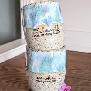 Könnte beinhalten: Zwei gestapelte Keramikbecher mit gesprenkelter beigefarbener Basis und blau-weiß glasierter Oberseite. Die Becher haben deutschen Text. Eine kleine rosa Blume liegt auf der Holzoberfläche neben den Bechern.