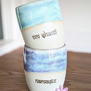 K&ouml;nnte beinhalten: Zwei gestapelte Keramikbecher mit hellblau-wei&szlig;er Glasur. Der obere Becher tr&auml;gt den Text "Om Shanti" und der untere Becher den Text "Namaste". Eine kleine rosa Blume liegt in der N&auml;he des unteren Bechers.