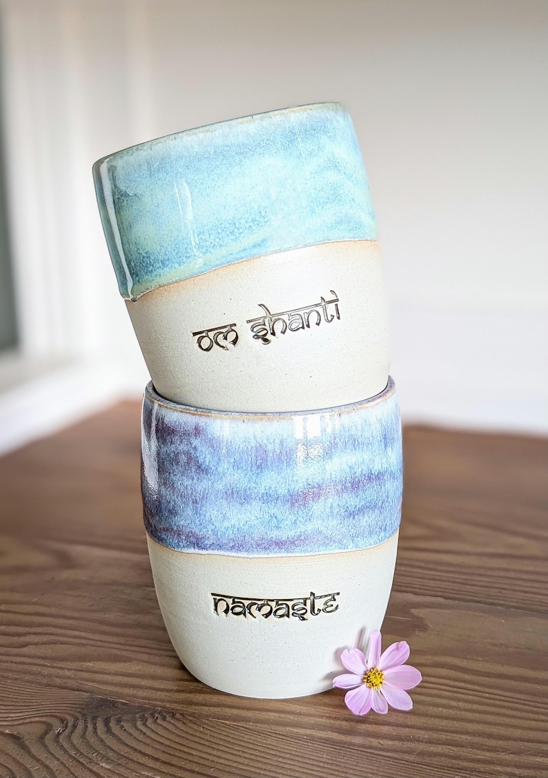 K&ouml;nnte beinhalten: Zwei gestapelte Keramikbecher mit hellblau-wei&szlig;er Glasur. Der obere Becher tr&auml;gt den Text "Om Shanti" und der untere Becher den Text "Namaste". Eine kleine rosa Blume liegt in der N&auml;he des unteren Bechers.