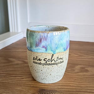 Könnte beinhalten: Ein Keramikbecher mit einer gesprenkelten cremefarbenen Basis und einer glänzenden blau-violett-grünen Tropfglasur am oberen Rand. Auf der Vorderseite steht der schwarze Text: wie schön, dass wir gleichzeitig leben. Der Becher ist zylindrisch.