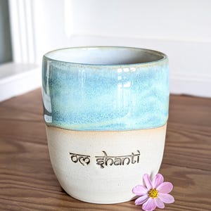 K&ouml;nnte beinhalten: Eine Keramik-Tasse mit hellblau-wei&szlig;er Glasur. Auf der Tasse steht der Schriftzug "Om Shanti" auf der unteren H&auml;lfte. Eine rosa Blume mit gelbem Zentrum liegt neben der Tasse auf einer Holzoberfl&auml;che.