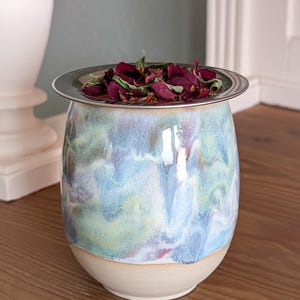 Könnte beinhalten: Eine Keramikvase mit blau, grün und weiß glasierter Oberfläche, gekrönt von einem silbernen Rand und gefüllt mit getrockneten Rosenblättern. Die Vase hat eine runde Form und einen beigen Sockel, der auf einer Holzoberfläche steht.