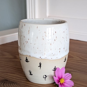 May include: A speckled white ceramic mug with a beige base. The base features black silhouettes of yoga poses. A pink flower with a yellow center rests beside the mug. The mug is on a wooden surface.