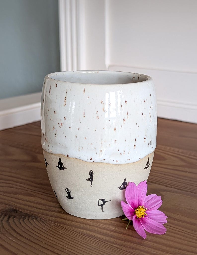 May include: A speckled white ceramic mug with a beige base. The base features black silhouettes of yoga poses. A pink flower with a yellow center rests beside the mug. The mug is on a wooden surface.