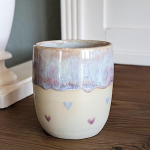 May include: A ceramic tumbler with a light beige base and a top section with a gradient of colors, including purple, blue, and tan. The base is decorated with small, heart-shaped designs in purple and blue. The tumbler sits on a wooden surface.