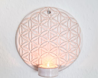 May include: White ceramic wall decor with a raised flower of life design and a candle holder. The candle holder is lit with a small tea light candle.