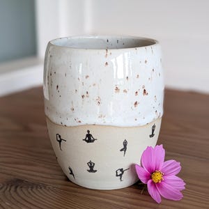 May include: A speckled white ceramic tumbler with a cream-colored base. The base features black yoga pose illustrations. A pink flower with a yellow center rests beside the tumbler. The tumbler is on a wooden surface.