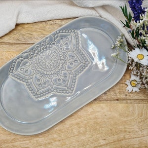Könnte beinhalten: Ein hellgraues ovales Serviertablett aus Keramik mit einem erhabenen, geprägten Mandala-Design. Das Tablett hat eine glänzende Oberfläche.