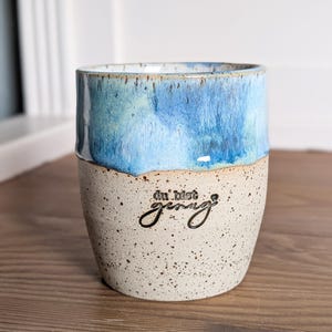 Könnte beinhalten: Ein Keramikbecher mit einer gesprenkelten cremefarbenen Basis und einem leuchtend blau glasierten oberen Rand. Der Text "du bist genug" ist in Schwarz auf der Vorderseite des Bechers eingeprägt. Das Design zeigt einen glatten Übergang.