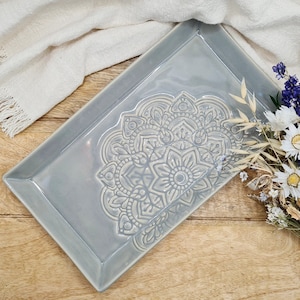 Könnte beinhalten: Ein rechteckiges Serviertablett aus hellgrauem Keramik mit einem geprägten Mandala-Muster. Das Tablett ist rechteckig und hat einen leicht erhöhten Rand.