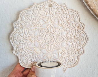 May include: White ceramic wall decor with a raised mandala design and a small candle holder attached. The candle holder has a white ceramic base and a small white candle inside.