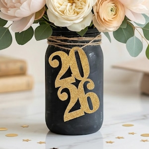 May include: A bouquet of blush pink peonies, cream roses, and eucalyptus leaves in a black mason jar. The jar is decorated with a gold glitter "2026" cutout, with a graduation cap in the background. Gold star confetti is scattered on the surface.