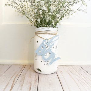May include: A white mason jar with a blue "It's a Boy" sign hanging from it. The jar is decorated with white paint and has a rustic look. The jar is filled with white baby's breath flowers.