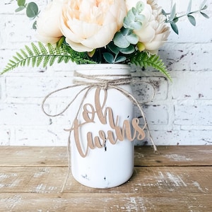May include: A white mason jar vase with a bouquet of peach-colored peonies, green ferns, and eucalyptus. The jar is adorned with a twine bow and a wooden cutout that reads "Oh Twins."