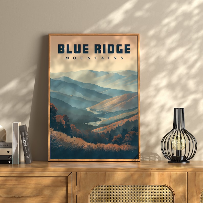 May include: A framed print with the text "BLUE RIDGE MOUNTAINS" above a landscape illustration. The artwork shows layered blue and brown mountain ranges, a river, and trees. The print is in a wooden frame and rests on a wooden cabinet.