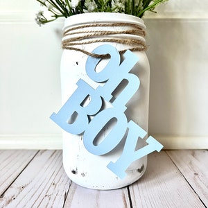 May include: White mason jar with a blue "It's a Boy" sign attached to it. The jar is decorated with twine and has a distressed white paint finish. The jar is filled with white baby's breath flowers.
