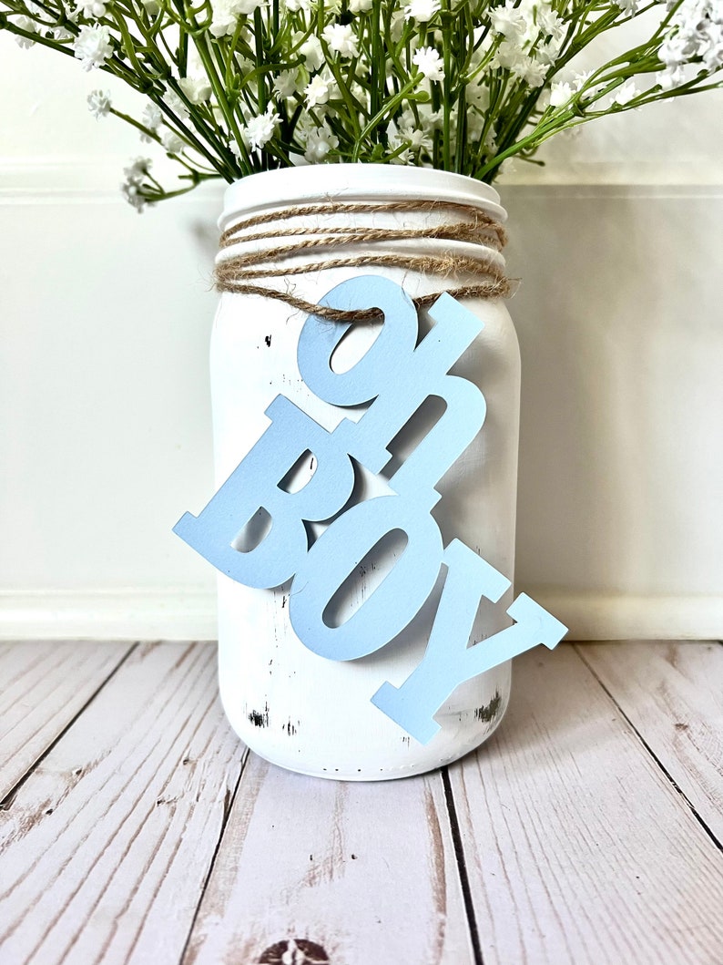 May include: White mason jar with a blue "It's a Boy" sign attached to it. The jar is decorated with twine and has a distressed white paint finish. The jar is filled with white baby's breath flowers.