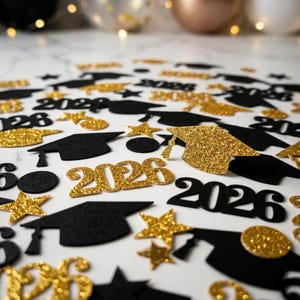 May include: A scattering of graduation-themed confetti in black and gold. The confetti includes mortarboard caps, stars, and the year "2026" in a glittery gold font. The background is a white surface with blurred decorative elements.