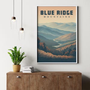 May include: A framed print depicting the Blue Ridge Mountains. The artwork shows a landscape of hills and a river, with the text "BLUE RIDGE MOUNTAINS" at the top. The colour scheme includes blues, greens, and earth tones, creating a serene mountain scene.
