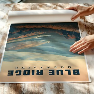 May include: A rolled poster featuring a landscape of the Blue Ridge Mountains. The artwork depicts a scenic view with blue and tan tones, with the words "BLUE RIDGE MOUNTAINS" printed at the bottom. The poster is being unrolled on a wooden surface.