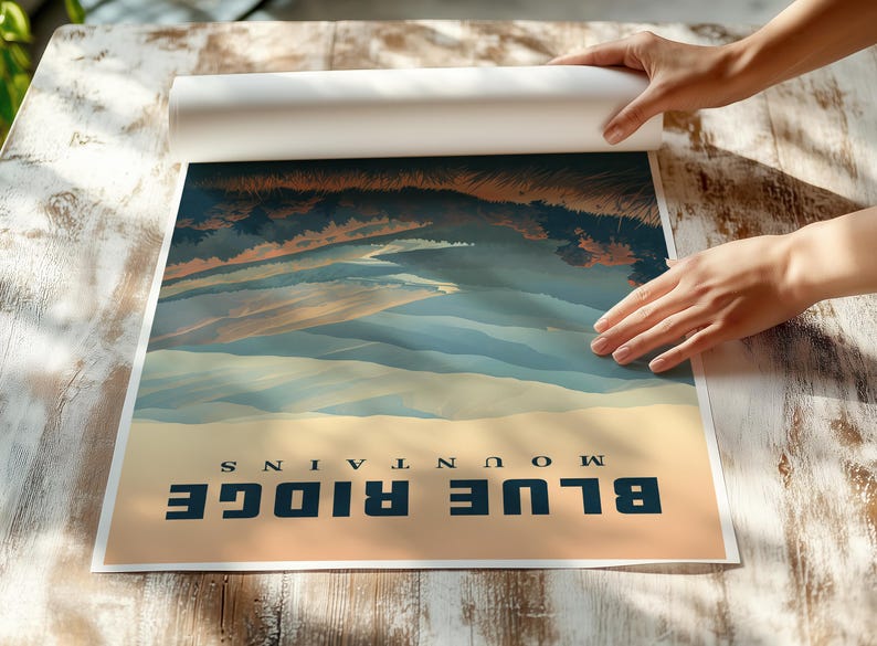 May include: A rolled poster featuring a landscape of the Blue Ridge Mountains. The artwork depicts a scenic view with blue and tan tones, with the words "BLUE RIDGE MOUNTAINS" printed at the bottom. The poster is being unrolled on a wooden surface.