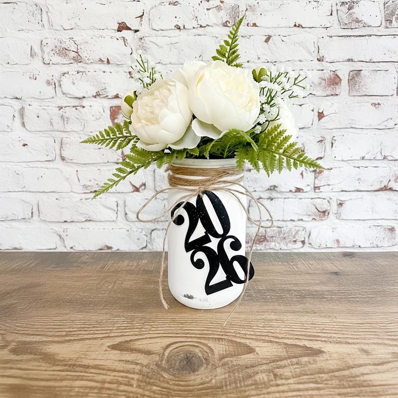 May include: A white ceramic mug with a black handle and the text "But first, coffee" in a handwritten style. The mug is on a white surface. The mug is approximately 4 inches tall.