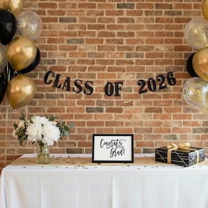 May include: A graduation celebration scene with black, gold, and clear balloons, a banner reading "CLASS OF 2026," and a sign that says "Congrats Grad!" A bouquet of white flowers and a wrapped gift box are on a table.