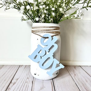 May include: A white mason jar with a blue "Oh Boy" sign hanging from the rim. The jar is decorated with twine and has a distressed white paint finish. The jar is filled with white baby's breath flowers.