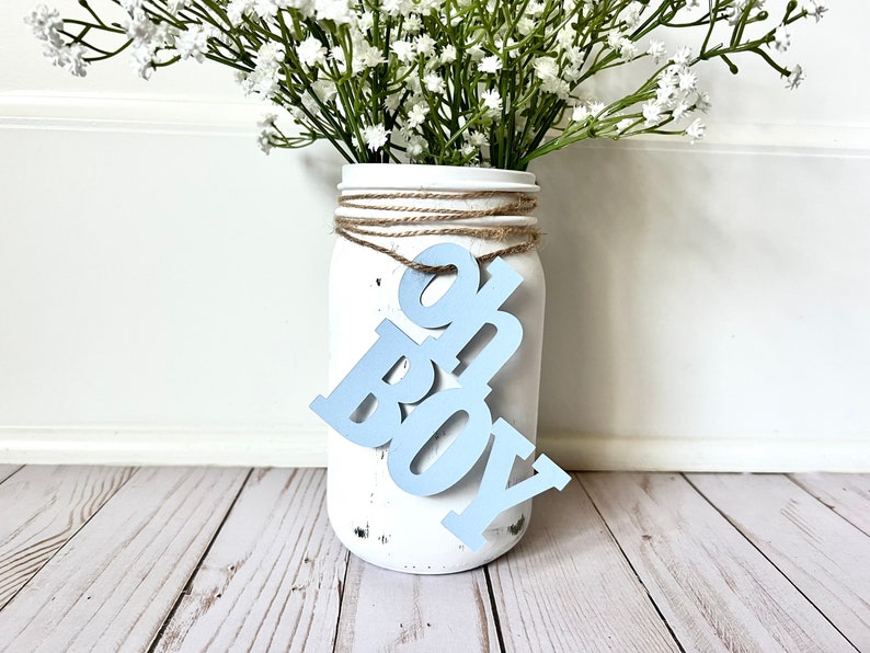 May include: A white mason jar with a blue "Oh Boy" sign hanging from the rim. The jar is decorated with twine and has a distressed white paint finish. The jar is filled with white baby's breath flowers.