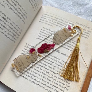 Pode incluir: Um marcador de p&aacute;gina dourado e branco com um design de flores prensadas e um tassel dourado. O marcador de p&aacute;gina &eacute; feito de resina e apresenta uma cita&ccedil;&atilde;o do livro "O Segredo" de Rhonda Byrne. A cita&ccedil;&atilde;o diz: "Veja as coisas que voc&ecirc; quer. Saiba que elas j&aacute; s&atilde;o suas. N&atilde;o pense nelas como suas, como pertencentes a voc&ecirc;, como j&aacute; em sua posse."