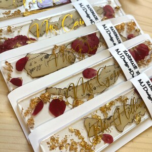Pode incluir: Conjunto de cinco marcadores de p&aacute;gina transparentes com folha de ouro e p&eacute;talas de flores vermelhas embutidas em resina. Cada marcador de p&aacute;gina tem um fundo branco e um nome escrito &agrave; m&atilde;o em letras douradas. Os nomes s&atilde;o "Sra. Cain", "Sra. Dominque", "Sra. Green", "Sra. Holloway" e "Sra. Smith".