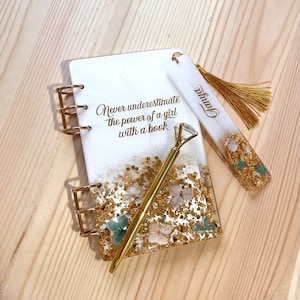 May include: A white journal with gold glitter and pressed flowers, a gold pen with a diamond-shaped top, and a white bookmark with gold glitter and pressed flowers. The journal has the quote "Never underestimate the power of a girl with a book" printed on the cover. The bookmark has the word "Jamie" written on it.
