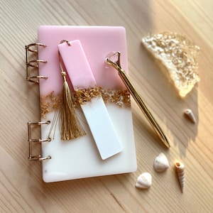 Peut inclure: Un carnet en résine rose et blanc avec des paillettes dorées et un marque-page assorti rose et blanc avec un pompon doré. Le carnet a une reliure spirale dorée et un stylo doré est posé à côté.