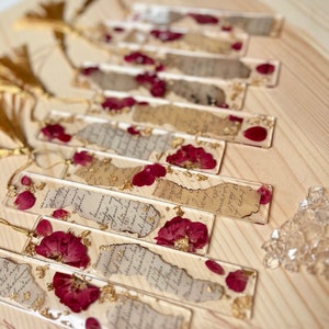 Peut inclure: Un ensemble de huit marque-pages faits à la main avec des fleurs séchées et des feuilles d'or enchâssées dans de la résine transparente. Chaque marque-page a un fond en papier vintage avec du texte manuscrit.