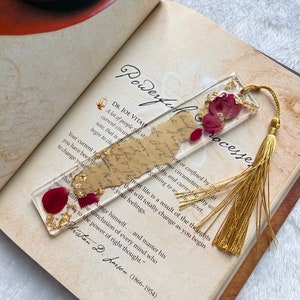 Pode incluir: Um marcador de p&aacute;gina dourado com uma borla vermelha e uma se&ccedil;&atilde;o de resina transparente contendo p&eacute;talas de rosa vermelha secas e folha de ouro. O marcador de p&aacute;gina est&aacute; sobre um livro com o texto "Powerful Processes" e uma cita&ccedil;&atilde;o de Christian D. Larson.