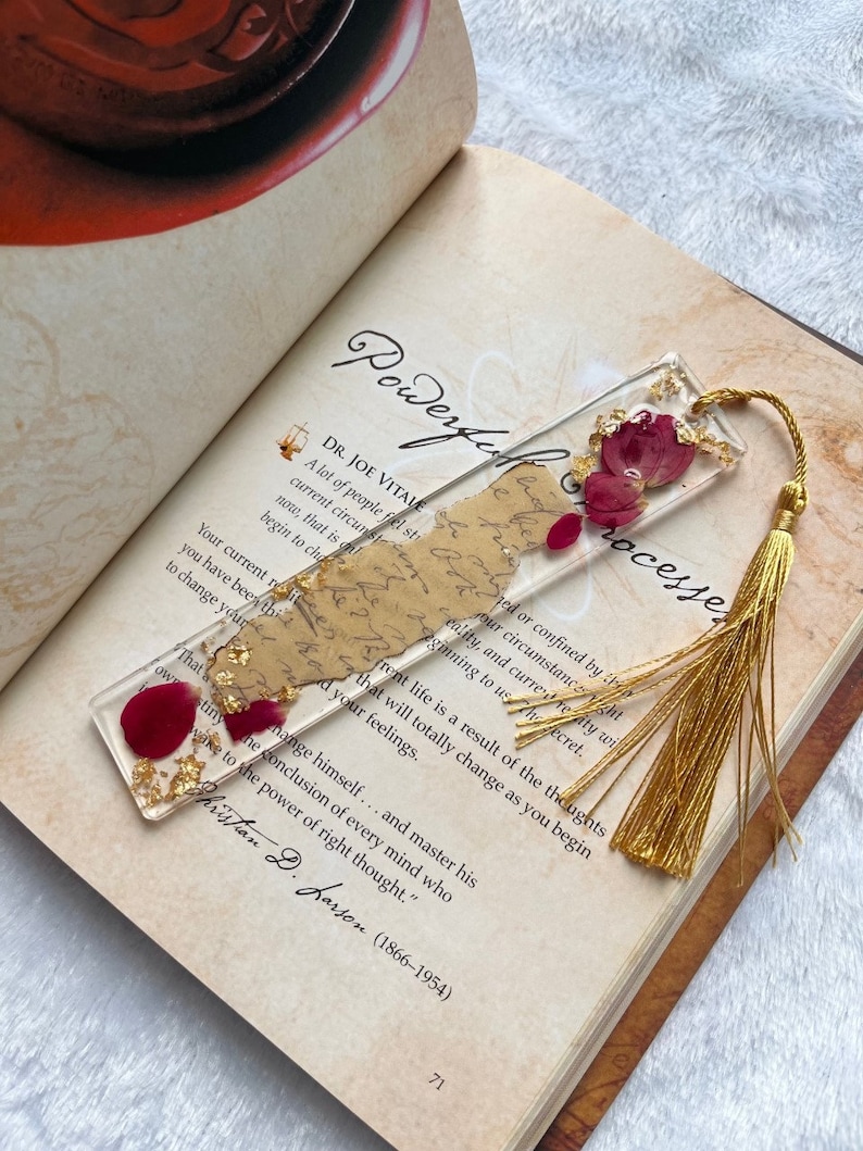 Pode incluir: Um marcador de p&aacute;gina dourado com uma borla vermelha e uma se&ccedil;&atilde;o de resina transparente contendo p&eacute;talas de rosa vermelha secas e folha de ouro. O marcador de p&aacute;gina est&aacute; sobre um livro com o texto "Powerful Processes" e uma cita&ccedil;&atilde;o de Christian D. Larson.