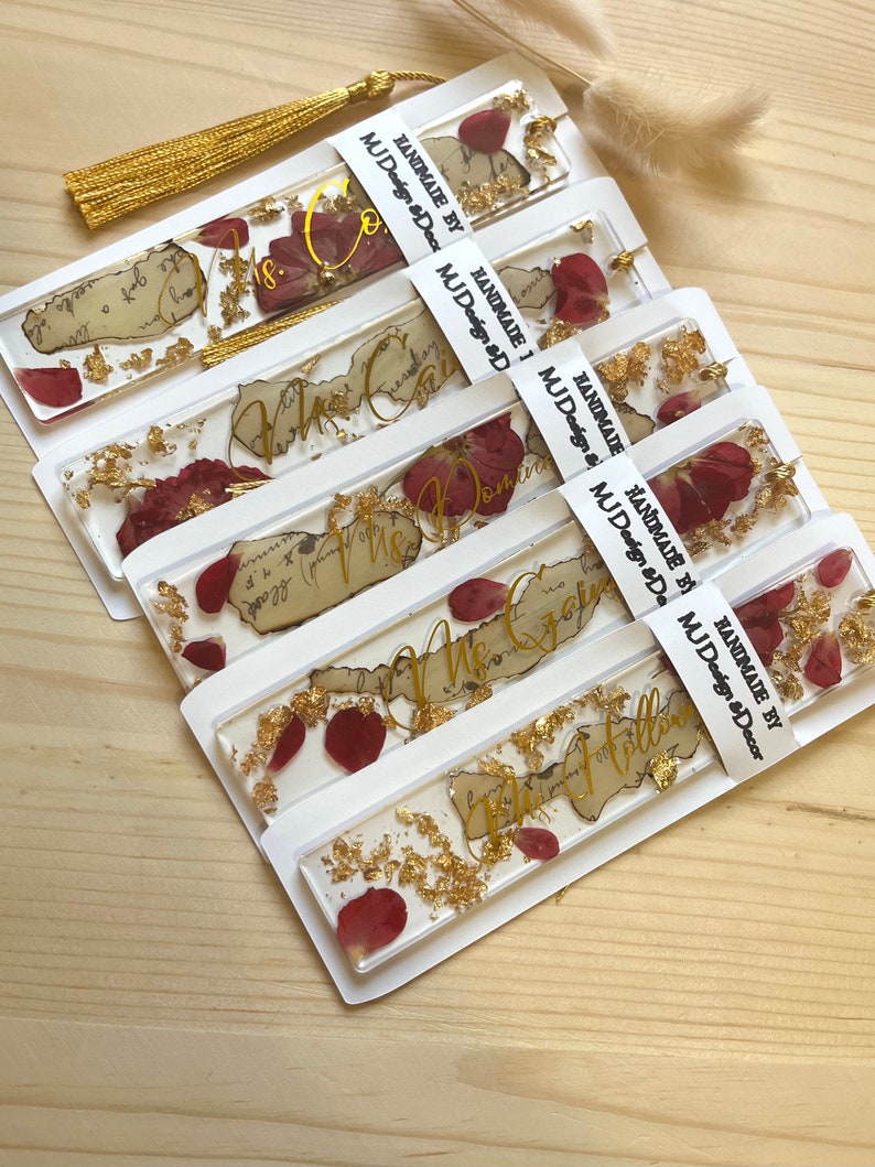 Pode incluir: Conjunto de seis marcadores de p&aacute;gina feitos &agrave; m&atilde;o com folha de ouro e p&eacute;talas de rosa secas embutidas em resina transparente. Cada marcador de p&aacute;gina apresenta um fundo de papel de estilo vintage com texto de caligrafia dourada. O texto nos marcadores de p&aacute;gina diz "The Calm", "The Domin", "The Gr", "The Holl", "The Follow", e "The Co".
