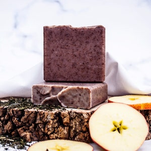 May include: Two rectangular, brown soaps with a speckled texture are stacked on a rustic wooden base. Sliced apples and green tea leaves are scattered around the base. The background is a white marble surface.