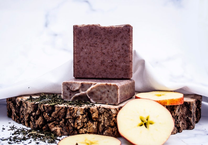 May include: Two rectangular, brown soaps with a speckled texture are stacked on a rustic wooden base. Sliced apples and green tea leaves are scattered around the base. The background is a white marble surface.