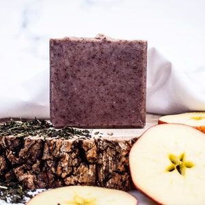 May include: A square, textured bar of soap with a speckled, dark brown appearance. The soap sits on a rustic wooden surface, accompanied by green tea leaves and apple slices. A white cloth is in the background.