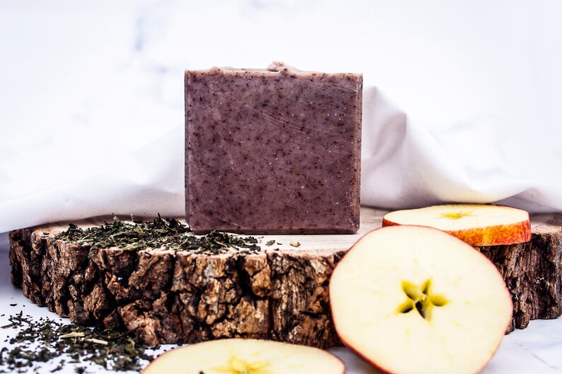 May include: A square, textured bar of soap with a speckled, dark brown appearance. The soap sits on a rustic wooden surface, accompanied by green tea leaves and apple slices. A white cloth is in the background.