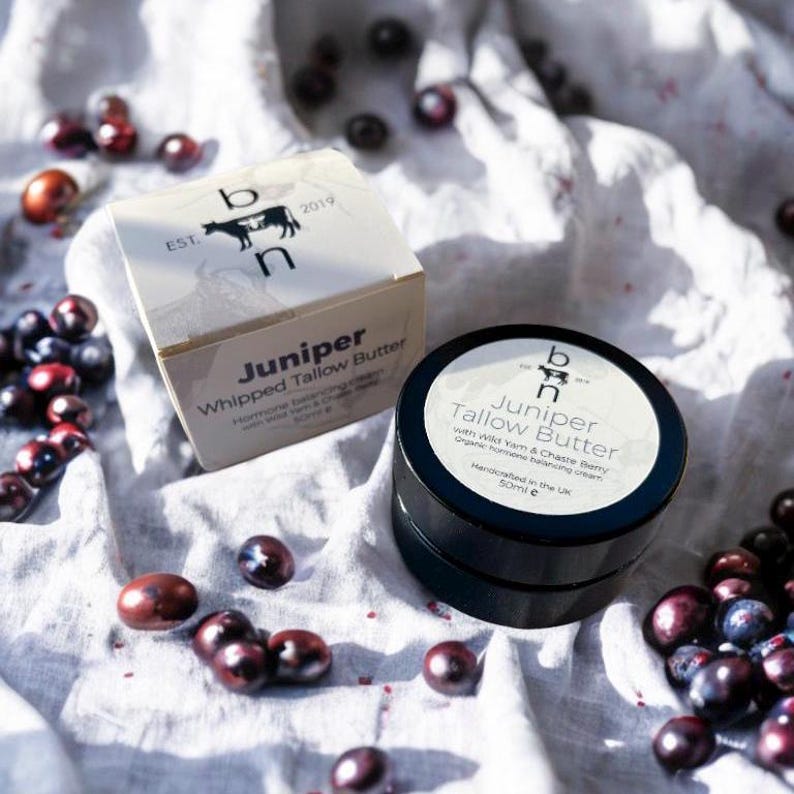 May include: A black jar of Juniper Whipped Tallow Butter with Wild Yam & Chaste Berry, organic hormone balancing cream. The jar is on a white fabric background with dark purple berries scattered around it. The jar is labeled with the brand name "b n" and the text "Handcrafted in the UK 50ml e".