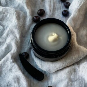 May include: A black jar with a white cream inside. The jar is sitting on a white fabric surface with dark brown coffee beans scattered around it. A black spatula is also in the image.