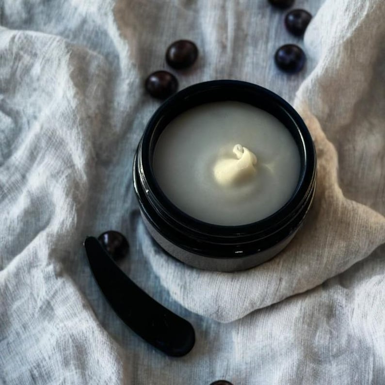 May include: A black jar with a white cream inside. The jar is sitting on a white fabric surface with dark brown coffee beans scattered around it. A black spatula is also in the image.