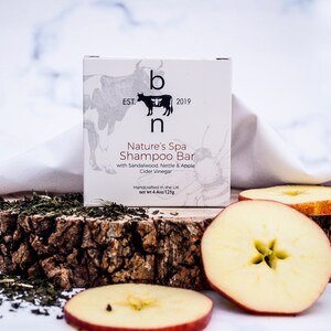 May include: A white box of Nature's Spa Shampoo Bar with the text "b n EST. 2019". The box is on a wooden slab with apple slices and green herbs. The shampoo bar contains sandalwood, nettle, apple cider vinegar. Net wt 4.4oz/125g.