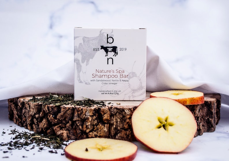 May include: A white box of Nature's Spa Shampoo Bar with the text "b n EST. 2019". The box is on a wooden slab with apple slices and green herbs. The shampoo bar contains sandalwood, nettle, apple cider vinegar. Net wt 4.4oz/125g.