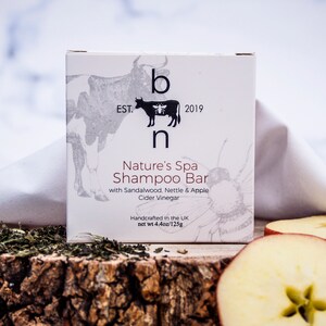 May include: A white box of Nature's Spa Shampoo Bar with a cow and bee logo, and the text "EST. 2019". The box is on a wooden surface with tea leaves and apple slices. The net weight is 4.4oz/125g.